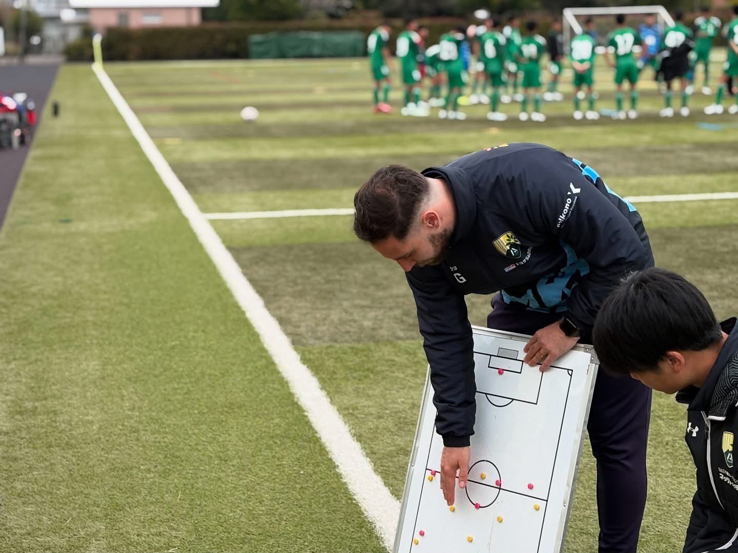 Pau Garnatje Font setting out a shape on the tactics board beside the pitch, players warming up behind